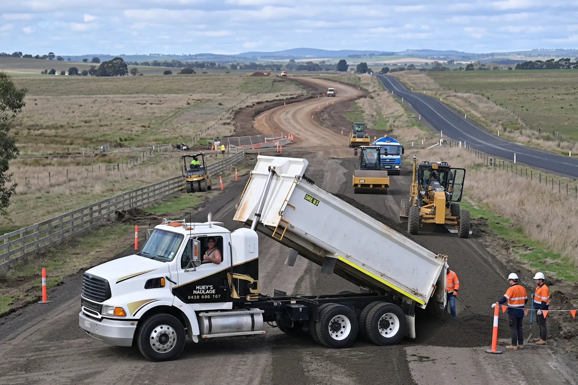 Huey's Haulage tipper truck unloading road base beside rollers and a grader on a rural road build